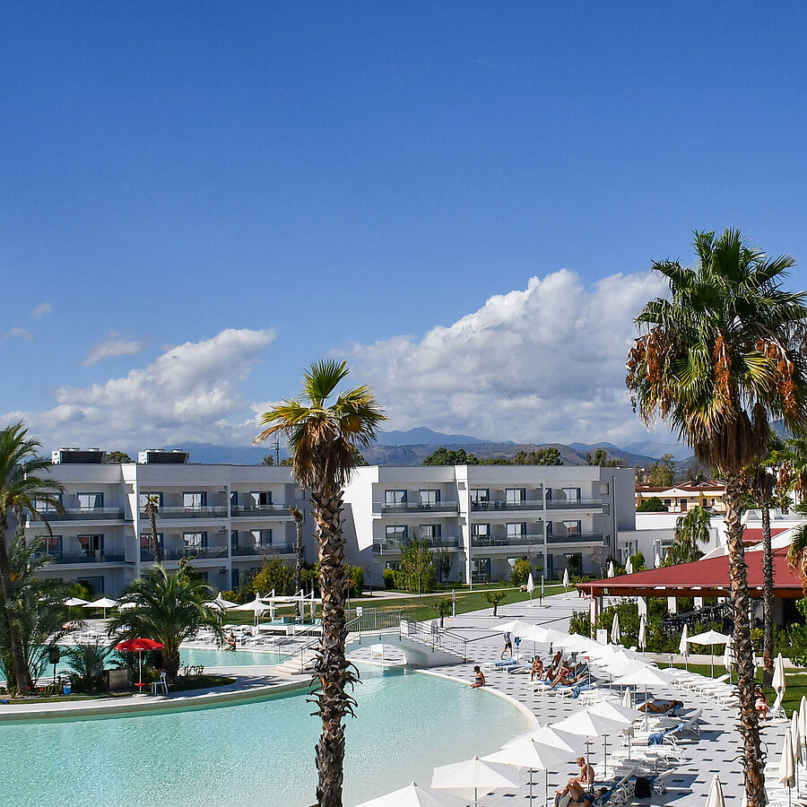 Außenansicht Panorama des Maritim Resort Calabria mit Pool, Palmen, weißen Gebäuden und Bergkulisse im Hintergrund bei klarem Himmel.