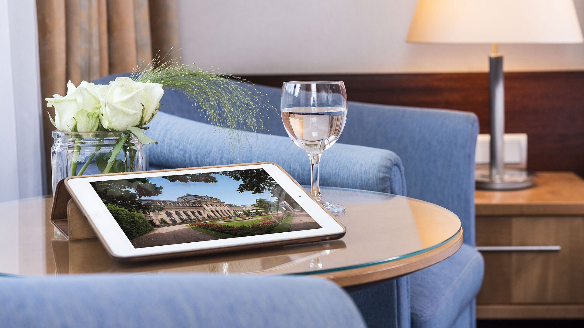 Accogliente area relax al Maritim Hotel Fulda con tablet, bicchiere d’acqua e rose bianche su tavolo in vetro.